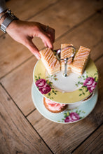 Load image into Gallery viewer, A hand is reaching down from the top left corner to take 1 of 3 layered tray bakes from the top plate of an Aynsley Archive Rose 2 Tier cake stand. The Plate on the bottom has a blue board with pink roses and green leaves. The plate on the top has a yellow border with pink roses and green leaves. The Cake stand pieces holding it together are silver in colour. It is sitting on a wooden table.