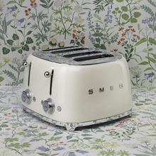 Load image into Gallery viewer, A floral background. Smeg 50's Retro Cream 4 Slice Toaster. The body of the toaster is Cream with chrome letters S, M, E and G embossed on either side. The top, base, levers, knobs and buttons are all chrome. There are two push down levers, two browning knobs, two defrost, two reheat and two stop buttons. One for each set of 2 slots.