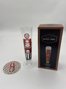 Tall slim Pilsner style beer glass wih red and black beer pump in the style of an american fuel pump. Matching checkered flag beer mat is to its left and gift box to its right.