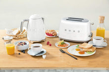 Load image into Gallery viewer, Lifestyle View. Smeg White 50s Retro 2 Slice Toaster and matching Kettle. The body of the toaster and kettle are white with chrome letters S, M, E and G embossed on both sides. The top and base of the toaster are chrome. The push down lever, defrost, reheat, stop buttons and browning knob on the toaster are all chrome. The handle, spout and base of the kettle are chrome. There are several various breakfast items sitting around the Smeg pieces to accessorize. Including: Bread, Sugar cubes and coffee.

