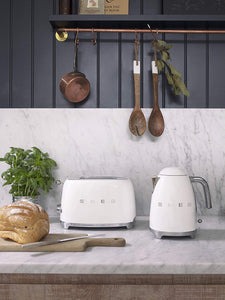 Lifestyle Image. Set in a kitchen with A copper pan and 2 wooden spoons hanging in the background. A potted plant and uncut bread sitting in the foreground. The Smeg 50s Retro White 2 Slice Toaster and matching Kettle are sitting in the middle on a grey marble worktop.