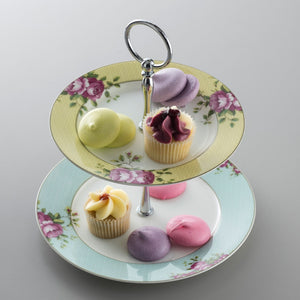 A grey background. This is an image of the Aynsley Archive Rose 2 Tier cake stand holding petit fours and lilac, lemon and pink coloured macarons. The tops plate has a yellow border. The bottom plate has a blue border. Both plates have pink rose and green leaf motifs. The pieces holding the cake stand together are silver in colour.
