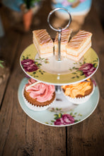 Load image into Gallery viewer, A lifestyle image of an Aynsley Archive Rose 2 tier cake stand. The Plate on the bottom has a blue board with pink roses and green leaves. The plate on the top has a yellow border with pink roses and green leaves. The Cake stand pieces holding it together are silver in colour. There are 3 layered traybakes on the top plate and 2 cupcakes on the bottom plate and it is sitting on a wooden table.
