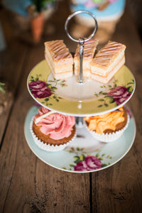 A lifestyle image of an Aynsley Archive Rose 2 tier cake stand. The Plate on the bottom has a blue board with pink roses and green leaves. The plate on the top has a yellow border with pink roses and green leaves. The Cake stand pieces holding it together are silver in colour. There are 3 layered traybakes on the top plate and 2 cupcakes on the bottom plate and it is sitting on a wooden table.