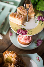 Load image into Gallery viewer, A close up of an Aynsley Archive Rose 2 tier cake stand. The Plate on the bottom has a blue board with pink roses and green leaves. The plate on the top has a yellow border with pink roses and green leaves. The Cake stand pieces holding it together are silver in colour. There are 3 layered traybakes on the top plate and a cupcake with pink icing on the bottom plate. There are coordinating items from the collection blurred into the background.
