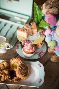 This is a lifestyle image of the Aynsley Archive rose collection sitting on a wooden table, accessorized with pastries, traybakes, buns and easter themed bunny figures holding coloured eggs and palm leaves.