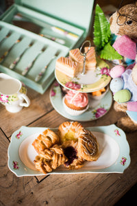 This is a lifestyle image of the Aynsley Archive rose collection sitting on a wooden table, accessorized with pastries, traybakes, buns and easter themed bunny figures holding coloured eggs and palm leaves.