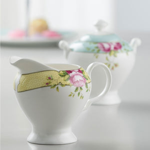 This is a close up of the cream jug on a white worktop, and the sugar bowl blurred into the background.  The Cream jug is white fine china with a yellow band around the rim. Decorated with delicate circles, pink roses and green leaves.