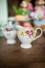 Load image into Gallery viewer, This is a lifestyle image of the Archive Rose cream jug on its own. Sitting on a wooden dining table, with matching items blurred into the background. The Cream jug is white fine china with a yellow band around the rim. Decorated with delicate circles, pink roses and green leaves.
