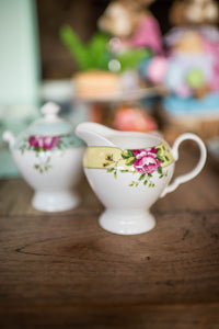 This is a lifestyle image of the Archive Rose cream jug on its own. Sitting on a wooden dining table, with matching items blurred into the background. The Cream jug is white fine china with a yellow band around the rim. Decorated with delicate circles, pink roses and green leaves.