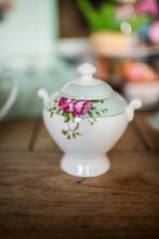Load image into Gallery viewer, This is a lifestyle images of the Archive Rose sugar bowl on its own. Sitting on a wooden dining table and a selection of matching items blurred into the background. The sugar bowl is white fine china with a blue band around with lid with delicate circles on the band. And a blue band around the rim of the bowl. There are pink roses and green leaves motifs on either side of the bowl, on the blue band. The bowl has 2 handles.
