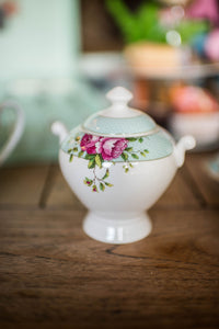 This is a lifestyle images of the Archive Rose sugar bowl on its own. Sitting on a wooden dining table and a selection of matching items blurred into the background. The sugar bowl is white fine china with a blue band around with lid with delicate circles on the band. And a blue band around the rim of the bowl. There are pink roses and green leaves motifs on either side of the bowl, on the blue band. The bowl has 2 handles.