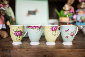 4 Aynsley Archive Rose footed fine china mugs in a row on a wooden dining table and an Aynsley gift box and Easter themed rabbits blurred into the background. 2 Yellow and 2 blue. All with pink roses, green leaves and a delicate circles all over the background colour. All handles are facing right.