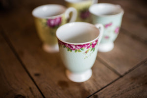 A close up of a blue footed mug on a wooden dining table and 3 other mugs from the set blurred into the background. This is a close up of the pink rose motif.