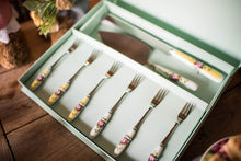 Load image into Gallery viewer, A close up of the set with a bokeh blur on the wooden dining table and co-ordinating archive rose pieces in the background. Aqua Display gift box. Contains 1 serrated blade cake knife with yellow, floral, fine china handle. 1 Stainless Steel Cake slice with blue floral fine china handle. 6 stainless Steel pastry forks. 3 hand blue floral fine china handles and 3 have yellow.
