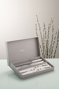 A Grey wall and a glass work top. There are willow branches in the background. The Aynsley Charbagh Pastry Set sits in its grey gift box. Stainless Steel with White Fine porcelain handles. A delicate persian and indian inspired motif on each handle. The set contains 1 cake slice, 1 serrated blade cake knife and 4 pastry forks.