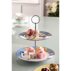 This is a lifestyle image of an Aynsley Cottage Garden 2 Tier cake stand. The both plates have a blue border. There are red and white roses, green leaves, blue and white daisies and blue butterflies in the pattern. The top plate is patterned all over. The bottom plate only has pattern around the border. The center of both plates is white. The pieces holding the cake stand up are silver in colour. In this picture the cake stand is sitting on a glass worktop and it is holding 6 petit fours.