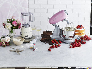Smeg 50s Retro Stand Mixer. The top of the mixer is Pink, the base is silver. There are the letters S, M, E and G embossed in chrome on either side of the machine. It has a Stainless Steel 4.8L bowl and beaters. The speed control is a chrome level on the top of the machine. The top lifts up by pressing in a button at the back of the motor case. Set beside a Smeg Cream Blender. Surrounded by cake, berries and flowers.