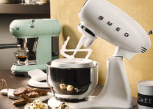 Load image into Gallery viewer, Smeg 50s Retro Stand Mixer. The body of the mixer is White. There are the letters S, M, E and G embossed in chrome on either side of the machine. It has a Stainless Steel 4.8L bowl and beaters. The speed control is a chrome level on the top of the machine. The top lifts up by pressing in a button at the back of the motor case. Sitting beside a Pastel Green Espresso Machine. Surrounded by chocolate biscuits and ice cream.
