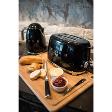 Load image into Gallery viewer, Lifestyle image in a kitchen with Black Smeg 50&#39;s Retro Kettle and 2 slice Toaster. They are sitting on a wooden chopping board with some bread, butter, jam and knives. 
