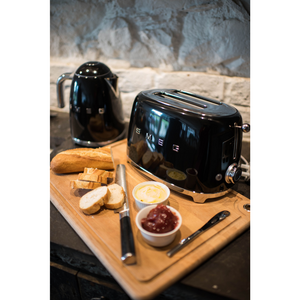 Lifestyle image in a kitchen with Black Smeg 50's Retro Kettle and 2 slice Toaster. They are sitting on a wooden chopping board with some bread, butter, jam and knives. 