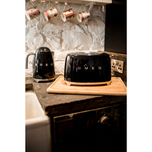 Load image into Gallery viewer, Lifestyle image in a kitchen with Black Smeg 50&#39;s Retro Kettle and 2 slice Toaster. There are 4 Julie Dodsworth mugs hanging from a shelf above the set. 
