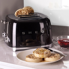Load image into Gallery viewer, Lifestyle Image. Set in a kitchen, on a grey marble worktop. The Smeg 50&#39;s Retro Black 4 Slice Toaster. The body of the toaster is black with chrome letters S, M, E and G embossed on either side. The top, base, levers, knobs and buttons are all chrome. There are two push down levers, two browning knobs, two defrost, two reheat and two stop buttons. One for each set of 2 slots. There are crumpets sitting up in the back 2 slots and on a white plate in the foreground. There is a cup of red herbal tea
