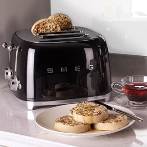 Lifestyle Image. Set in a kitchen, on a grey marble worktop. The Smeg 50's Retro Black 4 Slice Toaster. The body of the toaster is black with chrome letters S, M, E and G embossed on either side. The top, base, levers, knobs and buttons are all chrome. There are two push down levers, two browning knobs, two defrost, two reheat and two stop buttons. One for each set of 2 slots. There are crumpets sitting up in the back 2 slots and on a white plate in the foreground. There is a cup of red herbal tea