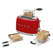 Load image into Gallery viewer, Lifestyle view. White Background. Smeg Red 50s Retro 2 Slice Toaster. The body of the toaster is red with chrome letters S, M, E and G embossed on both sides. The top and base are chrome. The push down lever, defrost, reheat, stop buttons and browning knob are all chrome. This picture shows the accessories available to purchase separately. A Toastie maker and a toast rack. These are filled with toasted wholemeal bread to demonstrate their purpose.
