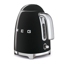 Load image into Gallery viewer, Rear side view. White Background. Smeg 50s Retro 1.7L Kettle. The body of the kettle is Black. There are chrome letters S, M, E and G embossed on each side. The lid is push button release. The spout, Handle, on/off lever and base are chrome. There is a water level window in line with the handle.
