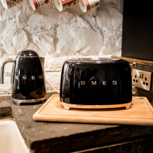 Lifestyle Image. In a kitchen setting on a black rustic worktop. White Brick wall Background. Smeg 50s Retro 1.7L Kettle. The body of the kettle is Black. There are chrome letters S, M, E and G embossed on each side. The lid is push button release. The spout, Handle, on/off lever and base are chrome. There is a water level window in line with the handle. Sitting next to the matching 2 slice toaster.