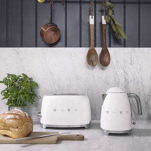 Lifestyle image. Set in a kitchen on a grey marble worktop. Smeg 50s Retro 1.7L Kettle and 2 Slice Toaster. The body of the kettle and toaster are Cream. There are chrome letters S, M, E and G embossed on each side. The lid is push button release. The spout, Handle, on/off lever and base are chrome. There is a water level window in line with the handle. The toaster has chrome slots, base, push lever, browning knob, reheat, defrost and stop buttons. 