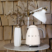 Load image into Gallery viewer, Lifestyle Image. Wooden shingle Background. Smeg 50s Retro 1.7L Kettle. The body of the kettle is Cream. There are chrome letters S, M, E and G embossed on each side. The lid is push button release. The spout, Handle, on/off lever and base are chrome. There is a water level window in line with the handle. It sits on a stone top table next to a white vase holding tan coloured leaves and in front of a White vintage desk lamp.
