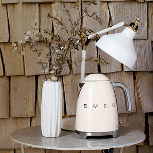 Lifestyle Image. Wooden shingle Background. Smeg 50s Retro 1.7L Kettle. The body of the kettle is Cream. There are chrome letters S, M, E and G embossed on each side. The lid is push button release. The spout, Handle, on/off lever and base are chrome. There is a water level window in line with the handle. It sits on a stone top table next to a white vase holding tan coloured leaves and in front of a White vintage desk lamp.