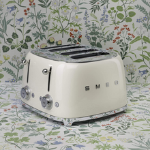 Load image into Gallery viewer, A floral background. Smeg 50&#39;s Retro Cream 4 Slice Toaster. The body of the toaster is Cream with chrome letters S, M, E and G embossed on either side. The top, base, levers, knobs and buttons are all chrome. There are two push down levers, two browning knobs, two defrost, two reheat and two stop buttons. One for each set of 2 slots.
