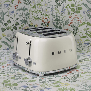 A floral background. Smeg 50's Retro Cream 4 Slice Toaster. The body of the toaster is Cream with chrome letters S, M, E and G embossed on either side. The top, base, levers, knobs and buttons are all chrome. There are two push down levers, two browning knobs, two defrost, two reheat and two stop buttons. One for each set of 2 slots.