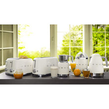 Load image into Gallery viewer, A lifestyle image of the Smeg 50s Retro collection in Cream. From left to right is: 4 Slice, 4 Slot Toaster. 4 Slice, 2 Slot toaster. Milk Frother. Citrus Juicer. Variable Temperature Kettle. They are sitting on a dark brown worktop in front of a window.
