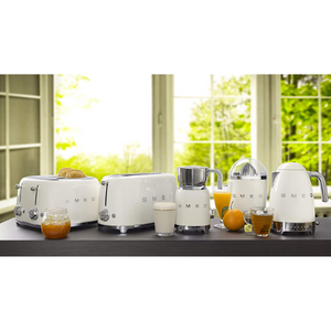 A lifestyle image of the Smeg 50s Retro collection in Cream. From left to right is: 4 Slice, 4 Slot Toaster. 4 Slice, 2 Slot toaster. Milk Frother. Citrus Juicer. Variable Temperature Kettle. They are sitting on a dark brown worktop in front of a window.