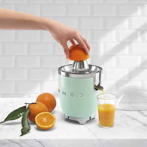 Lifestyle image in a white kitchen setting. A hand is pressing half an orange on to the reamer. Orange juice is pouring out of the spout into a glass. There are oranges and leaves to the left.