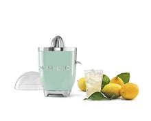 Load image into Gallery viewer, Lifestyle image. White background. The Smeg 50s Retro Citrus juicer. The body is pastel green. The reamer, spout and base are chrome. The clear dome that sits over the top of the reamer is sitting in the background. There are chrome letters S, M, E and G embossed on either side. There is a glass of juice with ice and 4 lemons with leaves to the right of the juicer.
