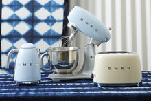 Load image into Gallery viewer, A lifestyle image of a Smeg Pastel Blue Kettle, Mixer and a Cream 2 Slice toaster. All sitting on a blue and white tie dye table cloth in front of a blue and whitetie dye backdrop. 
