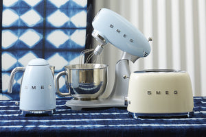 A lifestyle image of a Smeg Pastel Blue Kettle, Mixer and a Cream 2 Slice toaster. All sitting on a blue and white tie dye table cloth in front of a blue and whitetie dye backdrop. 